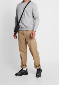 Sudadera gris con logo blanco, pantalones relajados de color beige y zapatillas negras con calcetines blancos. Bandolera negra colgada del hombro.