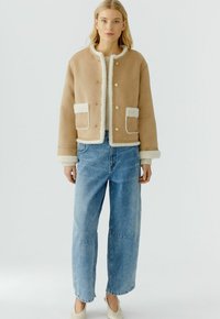 Beige cropped jack met nep-shearling afwerking en zakken, gouden knopen, gecombineerd met lichtblauwe wijde jeans en crèmekleurige schoenen.