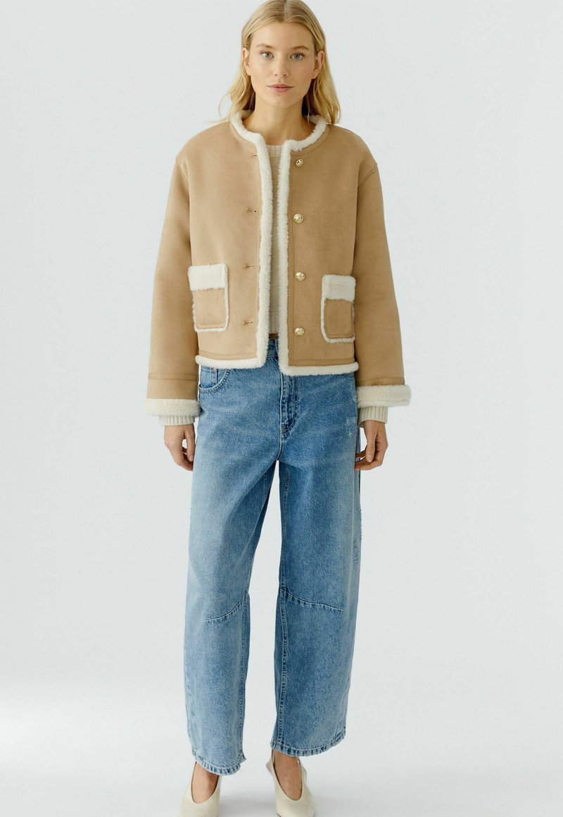 Beige cropped jack met nep-shearling afwerking en zakken, gouden knopen, gecombineerd met lichtblauwe wijde jeans en crèmekleurige schoenen.