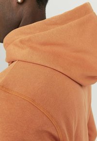 Sudadera naranja con una tela suave y texturizada, corte relajado y una gran capucha con costuras visibles y detalles de costura.