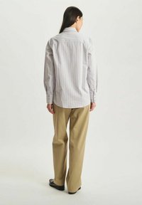 Chemise à manches longues blanche avec des fines rayures verticales rose et bleue. Elle présente un col pointu et des poignets boutonnés, assortie avec un pantalon beige.