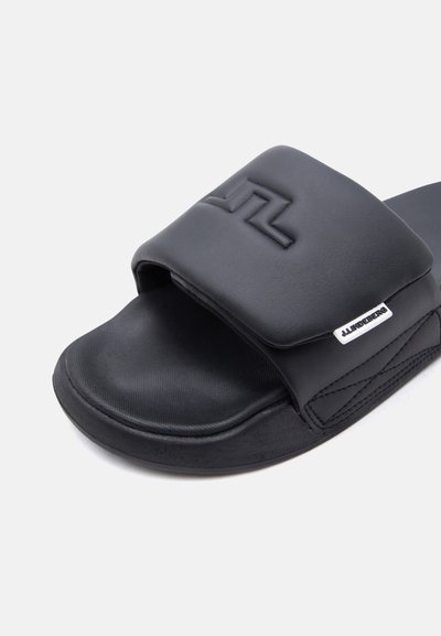 Μαύρο σανδάλι με φαρδιά επενδεδυμένη λουρίδα που διαθέτει ανάγλυφο λογότυπο και μικρή υφασμάτινη ετικέτα μάρκας στο πλάι της λουρίδας.