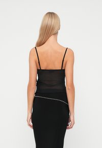 Camisole din plasă neagră cu bretele subțiri, purtat sub o fustă neagră ajustată, cu un accent din lanț argintiu și o textură netedă.