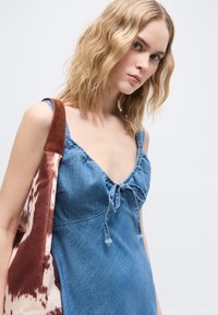 Ξανθιά γυναίκα με κυματιστά μαλλιά φορά μπλε μίνι φόρεμα από τζιν χωρίς μανίκια και κρατά καφέ και λευκή τσάντα ώμου με σχέδια, πάνω σε απλό φόντο.