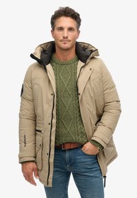 Chaqueta acolchada beige con capucha forrada de negro, mostrada sobre un suéter verde de punto trenzado. Las características incluyen bolsillos laterales y un logotipo en la manga.