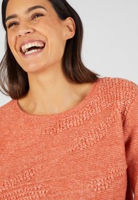 Pull en tricot de couleur corail vif avec des motifs texturés et un col rond large. Tissu doux avec une coupe décontractée.