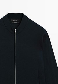 Giacca bomber di un intenso blu navy realizzata in 100% lana merino, con colletto a coste e chiusura con zip argentata, maniche lunghe e una texture liscia.