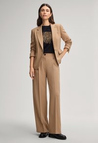 Un costume ajusté beige avec un pantalon large et un blazer cintré, porté sur un t-shirt noir avec un motif léopard. Chaussures noires.