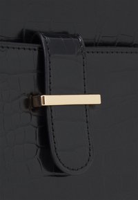 Nahaufnahme einer schwarzen, strukturierten Handtaschenklappe mit genähtem Riemen, der von einem horizontalen Metallverschluss in Goldton gesichert wird.