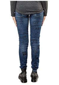 Los vaqueros ajustados de mezclilla azul oscuro cuentan con varios gráficos y textos en relieve. El diseño incluye bolsillos y una silueta ajustada. Se complementan con botas negras.