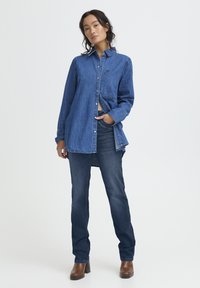 Donna che indossa una camicia di jeans blu a maniche lunghe, jeans blu scuro e stivali marroni con tacco, in piedi con una mano in tasca su sfondo bianco.