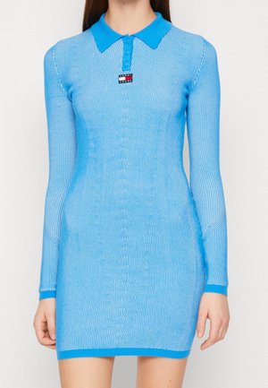 Kobieta ubrana w dopasowaną, długorękawową, niebieską prążkowaną dzianinową mini sukienkę z kołnierzykiem i logo Tommy Jeans na piersi.