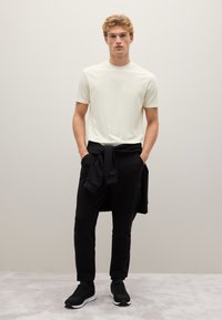 Witte t-shirt met korte mouwen, gecombineerd met zwarte joggers, en een zwart sweatshirt dat om de taille is gestrikt, met zwarte sneakers. Eenvoudig, strak ontwerp.