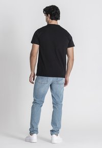 Čierne tričko s krátkym rukávom v kombinácii so svetlomodrými džínsami, s voľným strihom a bielymi teniskami. Model je zobrazený zozadu.