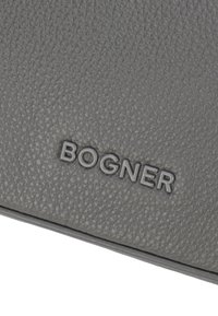 Šedý texturovaný kožený povrch s reliéfní značkou "BOGNER" v kontrastní tmavší barvě.
