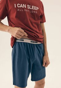 Czerwony T-shirt z białym napisem "MOGĘ SPAĆ CAŁE DNI" w połączeniu z granatowymi szortami z czarnym pasem oznaczonym "HENDERSON."