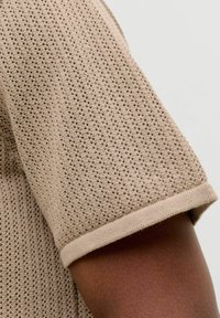 Haut en maille beige à manches courtes avec petit motif perforé porté sur un bras à peau foncée sur fond uni.
