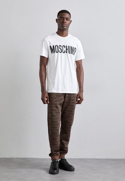 Balta medvilninė marškinėliai su juodu užrašu "MOSCHINO", derinami su rudos spalvos kelnėmis, kurių audinyje išspausdintas pakartotinis logotipas, dėvimi su juodais sportbačiais.
