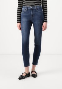 High-waisted, skinny blue jeans with a smooth texture and subtle fading. Paired with black flats featuring eyelet accents and a pointed toe.