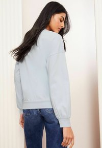 Felpa oversize blu chiaro con maniche a palloncino, texture morbida e polsini elasticizzati, abbinata a jeans blu scuro, vista da dietro.