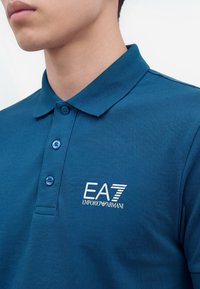 Homme portant un polo bleu à trois boutons avec un logo blanc EA7 Emporio Armani sur la poitrine.