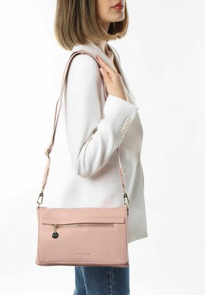 Roze leren crossbody tas met een voorzak met rits, gouden hardware-accenten en een verstelbare schouderriem, gedragen over een witte blazer en blauwe jeans.