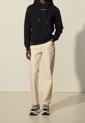Person trägt einen schwarzen Hoodie, cremefarbene Hosen und mehrfarbige Sneaker und steht vor einem schlichten hellen Hintergrund.