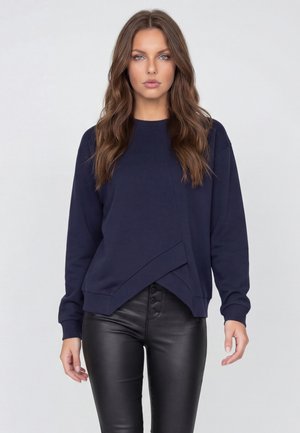 Frau mit langen, welligen braunen Haaren, die einen marineblauen asymmetrischen Pullover und schwarze, mit Knöpfen versehene Lederhosen trägt, steht vor einem schlichten Hintergrund.