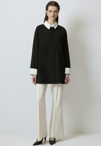 Zwarte oversized blouse met een witte kraag, knoopsluiting en manchetaccenten. Gecombineerd met crèmekleurige wijde broeken en zwarte puntige hakken.