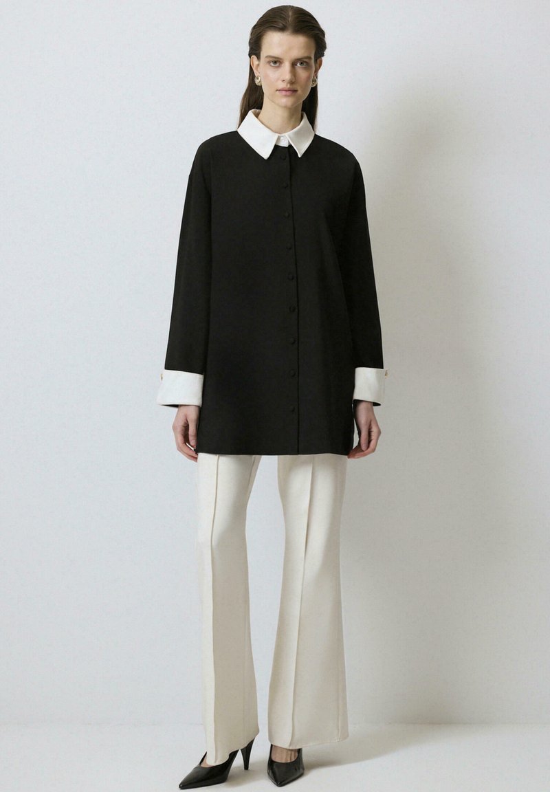 Zwarte oversized blouse met een witte kraag, knoopsluiting en manchetaccenten. Gecombineerd met crèmekleurige wijde broeken en zwarte puntige hakken.