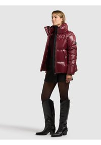 Bordeauxrode donzen jas met een hoge kraag, glanzend materiaal en ritssluitingen aan de zakken, gecombineerd met een zwarte rok en zwarte laarzen tot op de knie.