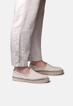 Persona che indossa espadrillas beige senza lacci con suole di corda e pantaloni larghi di colore chiaro, in piedi su uno sfondo bianco.