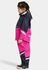 Regenpak bestaande uit een marineblauwe en roze waterdichte jas en bijpassende broek, met reflecterende strepen en elastische manchetten, gecombineerd met zwarte laarzen.
