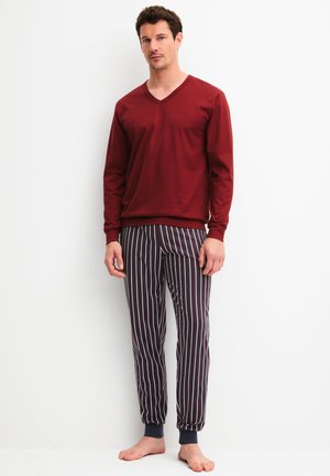 Man draagt een bordeauxrode, lange mouwen V-hals shirt en zwart-witte verticale gestreepte broek, staat blootsvoets tegen een witte muur.