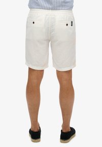 Weiße Leinen-Shorts mit strukturiertem Finish, ausgestattet mit zwei hinteren Taschen mit braunen Knöpfen und umgeschlagenen Säumen. Getragen mit schwarzen Slip-On-Schuhen.