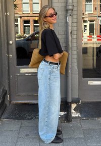 Svart kortärmad t-shirt, ljusblå baggy jeans, vävd beige handväska, svart bälte, runda solglasögon, rakt blont hår, stående vid en dörr.