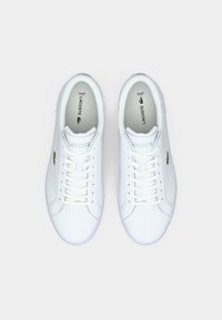 Paire de baskets blanches Lacoste avec lacets, logo crocodile vert sur les côtés, et semelles intérieures marquées, vues d'en haut sur un fond blanc.