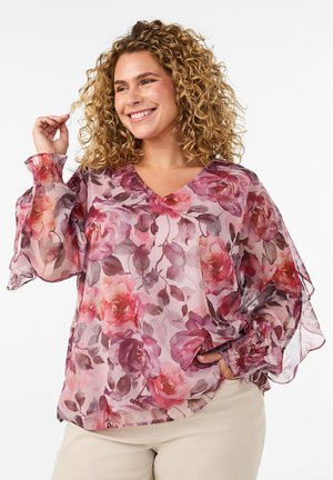 Frau mit lockigem blondem Haar, die eine durchsichtige, florale rosa-lila Bluse mit langen, aufgepufften Ärmeln und beiger Hose trägt, lächelnd und posierend.