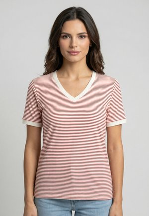 Jonge vrouw met donker haar draagt een rood-wit gestreept T-shirt met korte mouwen en V-hals, en lichtblauwe spijkerbroek, staand tegen een effen achtergrond.