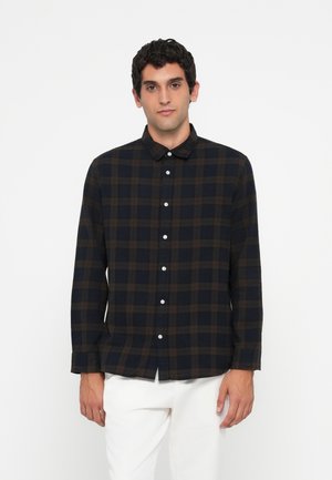 Young man with short dark hair wearing a black and brown plaid button-up shirt and white pants against a plain white background.
