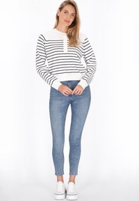 Nainen seisoo itsevarmana raidallisessa collegepaidassa ja kapeissa farkuissa, kädet ristissä, casual-valkoiset lenkkarit jalkoillaan yksinkertaista taustaa vasten.