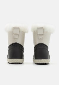 Las botas presentan un upper sintético beige con forro de piel sintética, base de goma negra y una suela con agarre texturizado. Incluyen un bucle en la parte trasera para facilitar su colocación.