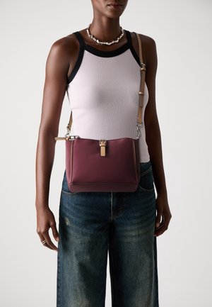 Femme portant un haut sans manches rose pâle avec un bord noir, un jean bleu et un sac bandoulière bordeaux avec une sangle beige traversant son torse.