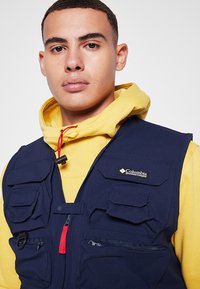 Gilet Columbia blu navy con multiple tasche frontali, una cerniera rossa e tessuto testurizzato, indossato sopra una felpa gialla con cappuccio.