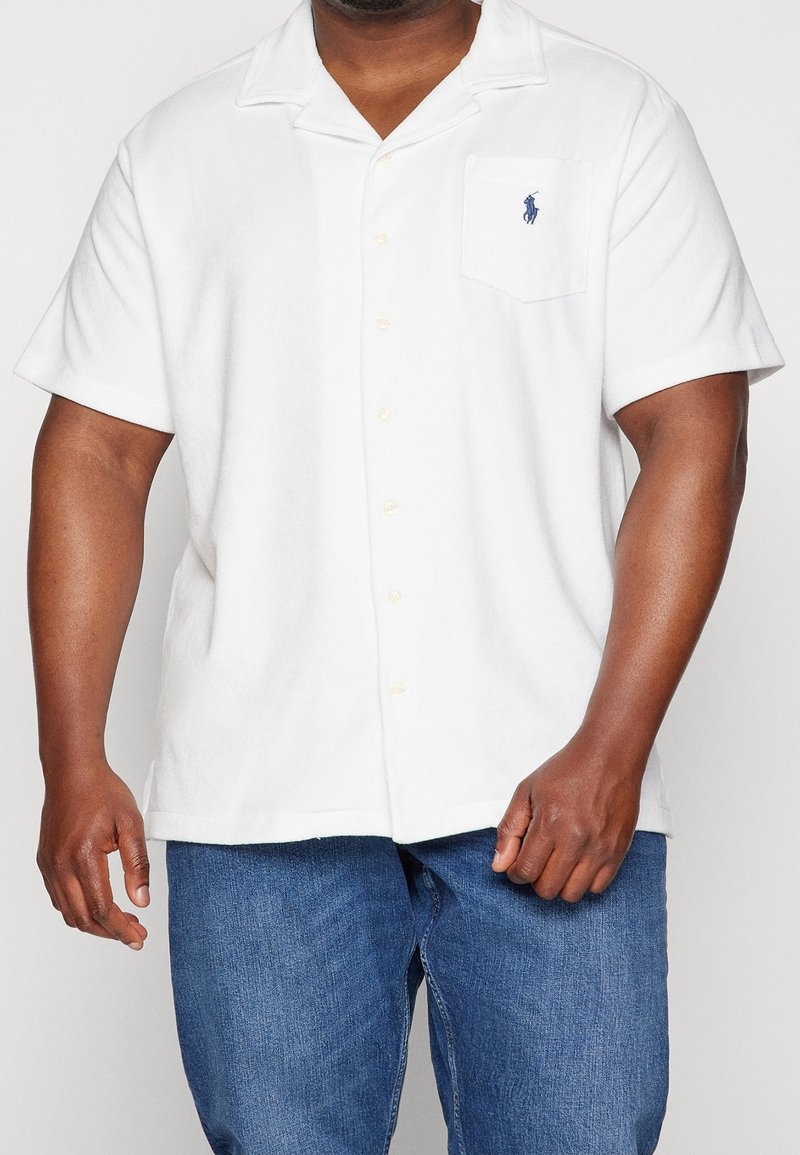 Hombre vistiendo una camisa blanca de manga corta con un logo en el bolsillo del pecho y unos jeans azules, sobre un fondo sencillo.