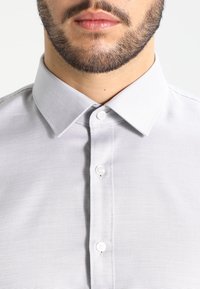 Chemise à boutons gris clair avec un col, tissu texturé et détail de boutons visible sur le devant et le col.