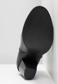 Sort lædersko med blokhæle, der hviler på en hvid overflade, sålen vender mod kameraet mod en hvid hjørnebagt.