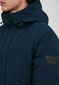 Navy blue winter jacket with a high collar and adjustable hood, featuring a textured patch on the sleeve and diagonal stitching details.