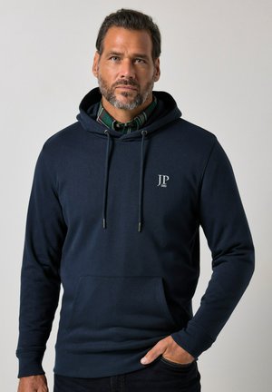 Man met baard die een marineblauwe hoodie draagt over een groen geruit overhemd, staand tegen een grijze achtergrond.
