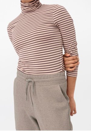 Osoba ubrana w beżowo-brązowy golf w paski oraz jasnoszare spodnie dresowe z troczkami, trzymająca jedną ręką za przeciwne ramię.